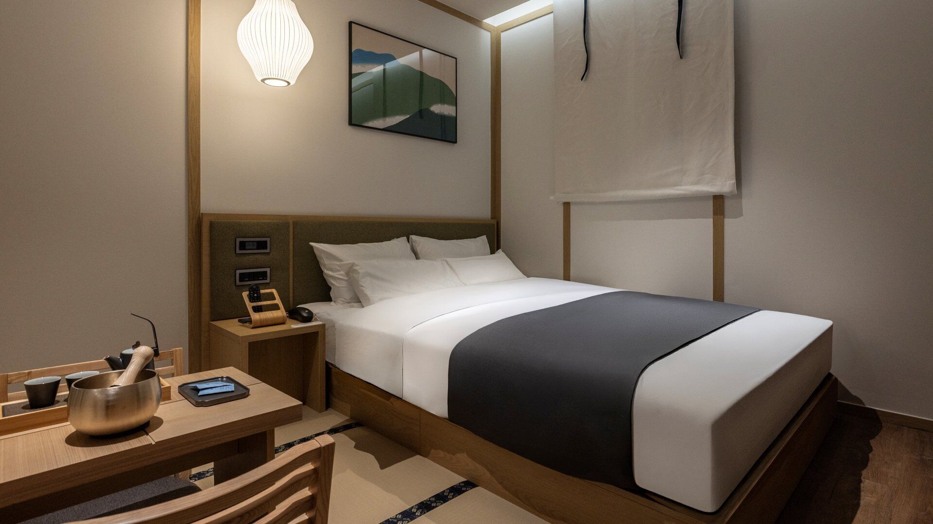 A hotel room with a bed, bedside table, lamp, and a painting on the wall.
