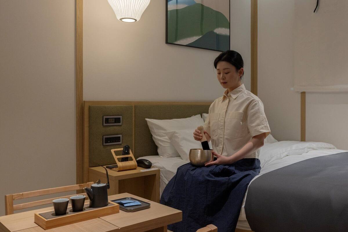 A hotel room with a bed, bedside table with a kettle, cups, and a smartphone.