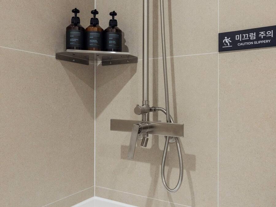 Zona de ducha con repisa metálica que contiene tres botellas, un letrero de precaución y un cabezal de ducha.