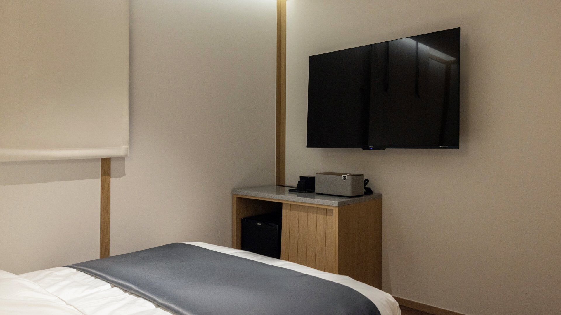 A hotel room with a bed, a TV mounted on the wall, a small wooden nightstand, and a built-in mini-fridge.