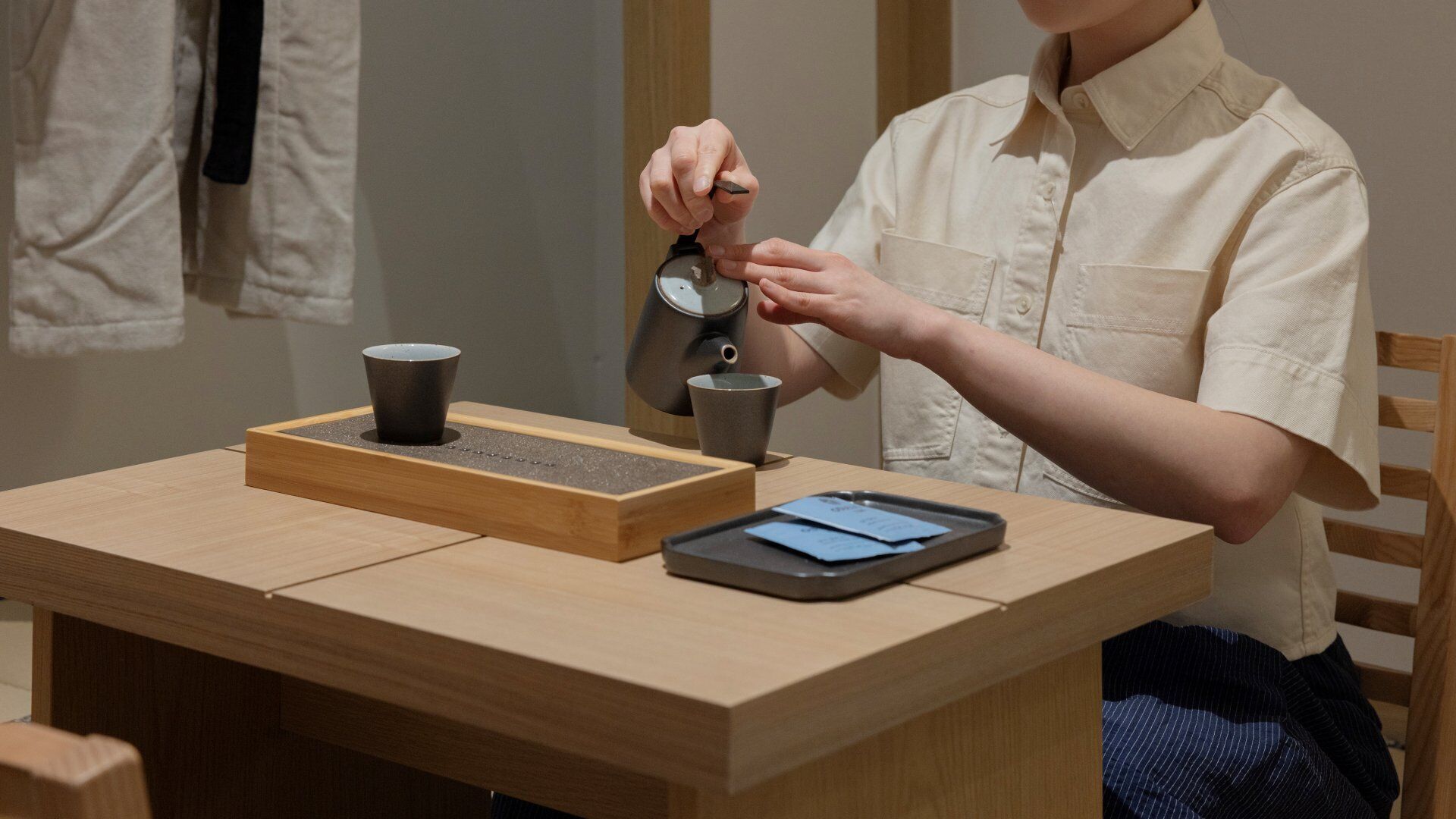 A person sitting at a wooden table with a cup, a mug, and a tray with a card.