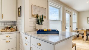A kitchen with white cabinets, gold handles, a marble countertop, a potted plant, and a dining area with a table and chairs. - Downtown Redmond 4BR Walk to Food & Brews (Redmond)