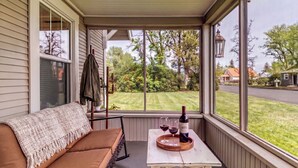 A covered porch with a cushioned seating area, a table with a bottle and glasses, and a view of a residential street and greenery. - Downtown Redmond 4BR Walk to Food & Brews (Redmond)