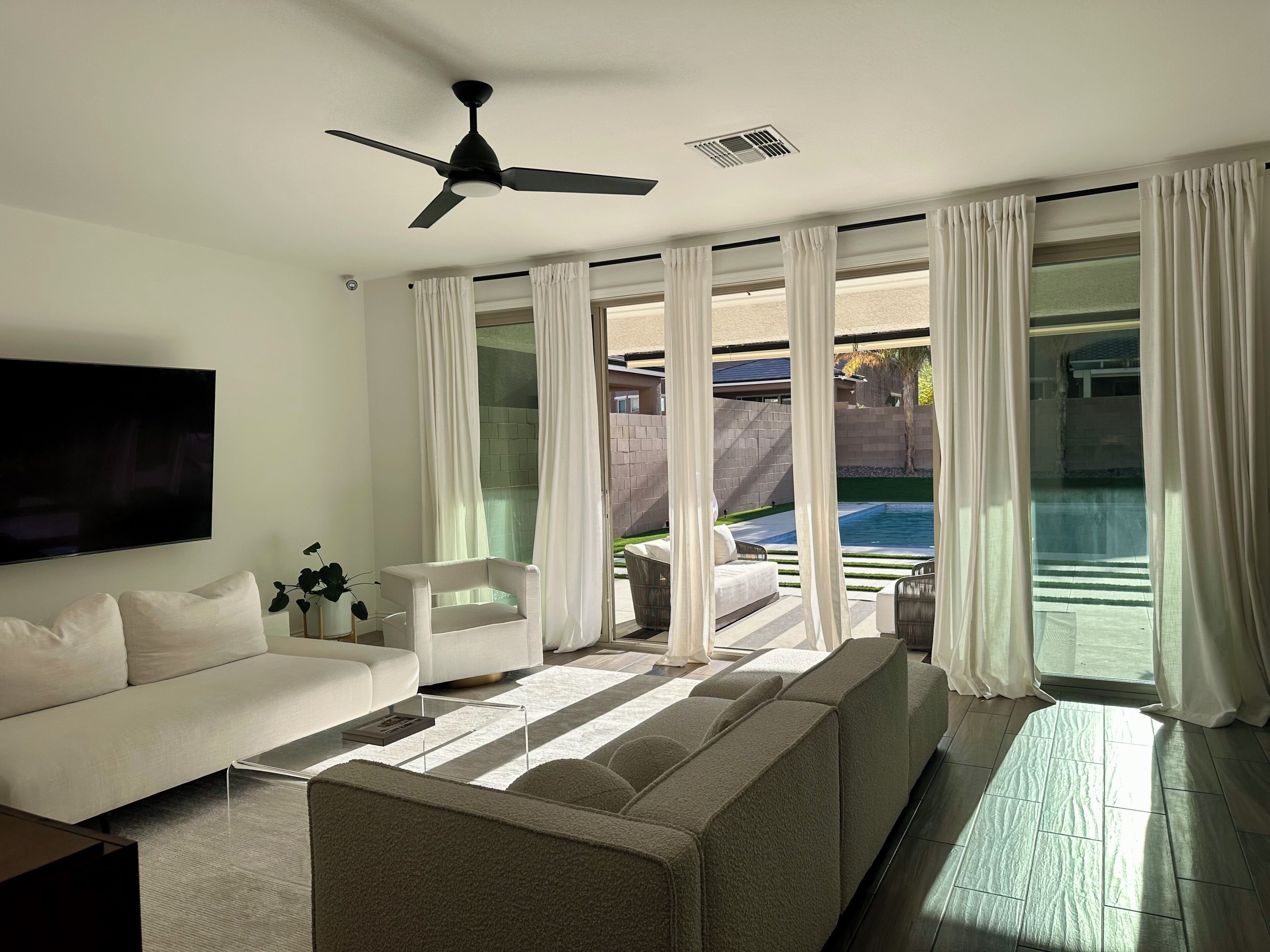 Un salon moderne avec un canapé, un téléviseur, un ventilateur de plafond et de grandes fenêtres dotées de rideaux.