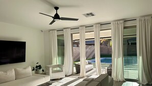 A modern living room with a sofa, a TV, a ceiling fan, and large windows with curtains. - Luxury Modern Retreat with Pool & Spa – Near the Strip (Las Vegas)