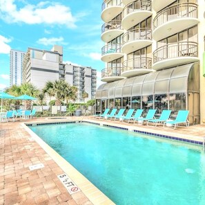 A swimming pool with lounge chairs, a glass-enclosed structure, and a multi-story building with balconies. - Myrtle Beach Oceanfront Escape w/Private Balcony - Stunning Beach & City Views (Myrtle Beach)