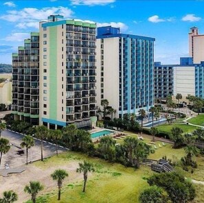 A multi-story hotel with balconies, a swimming pool, and a landscaped area with palm trees. - Myrtle Beach Oceanfront Escape w/Private Balcony - Stunning Beach & City Views (Myrtle Beach)