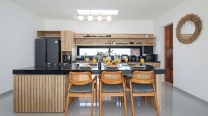 A modern kitchen with a central island, wooden chairs, and a dining table set for four. - Villa Cansavina (Seminyak)