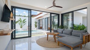 A modern living room with a large grey sofa, a round wooden coffee table, and a glass door leading to a pool area. - Villa Cansavina (Seminyak)