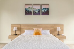 Una camera d'albergo con una testiera in legno, un letto grande con biancheria da letto bianca, due comodini con lampade e tre quadri con paesaggi appesi al muro.