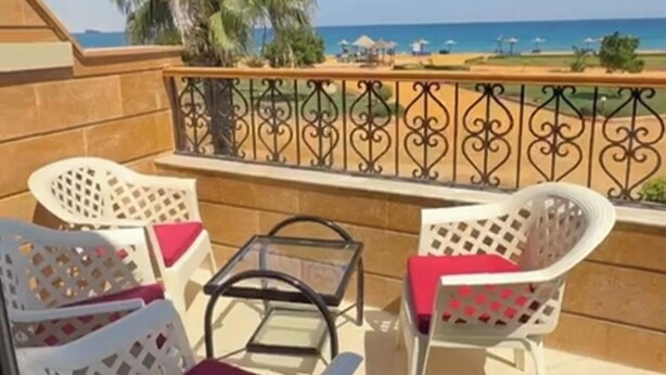 Un balcon avec des chaises en plastique blanches et une petite table, donnant sur une plage bordée de palmiers et sur l’océan.
