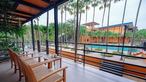 A covered outdoor seating area with wooden benches, overlooking a pool and a building with orange walls and blue roof. - Nongnooch Resort Pattaya (Sattahip)