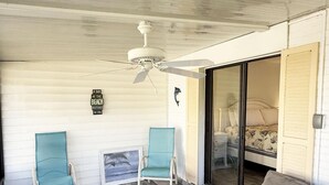 Une pièce dotée d’un ventilateur de plafond, de deux chaises, d’un canapé, d’un lit recouvert d’une courtepointe à motifs floraux et d’une porte-fenêtre coulissante donnant sur une autre pièce.