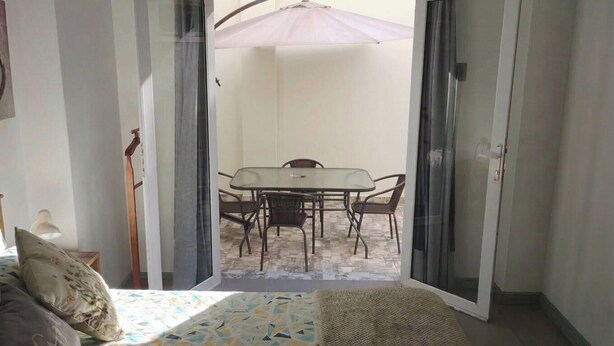 Eine offene Schiebetür führt zu einer Terrasse mit Tisch und Stühlen, einem Bett mit gemustertem Überwurf und einer Lampe.