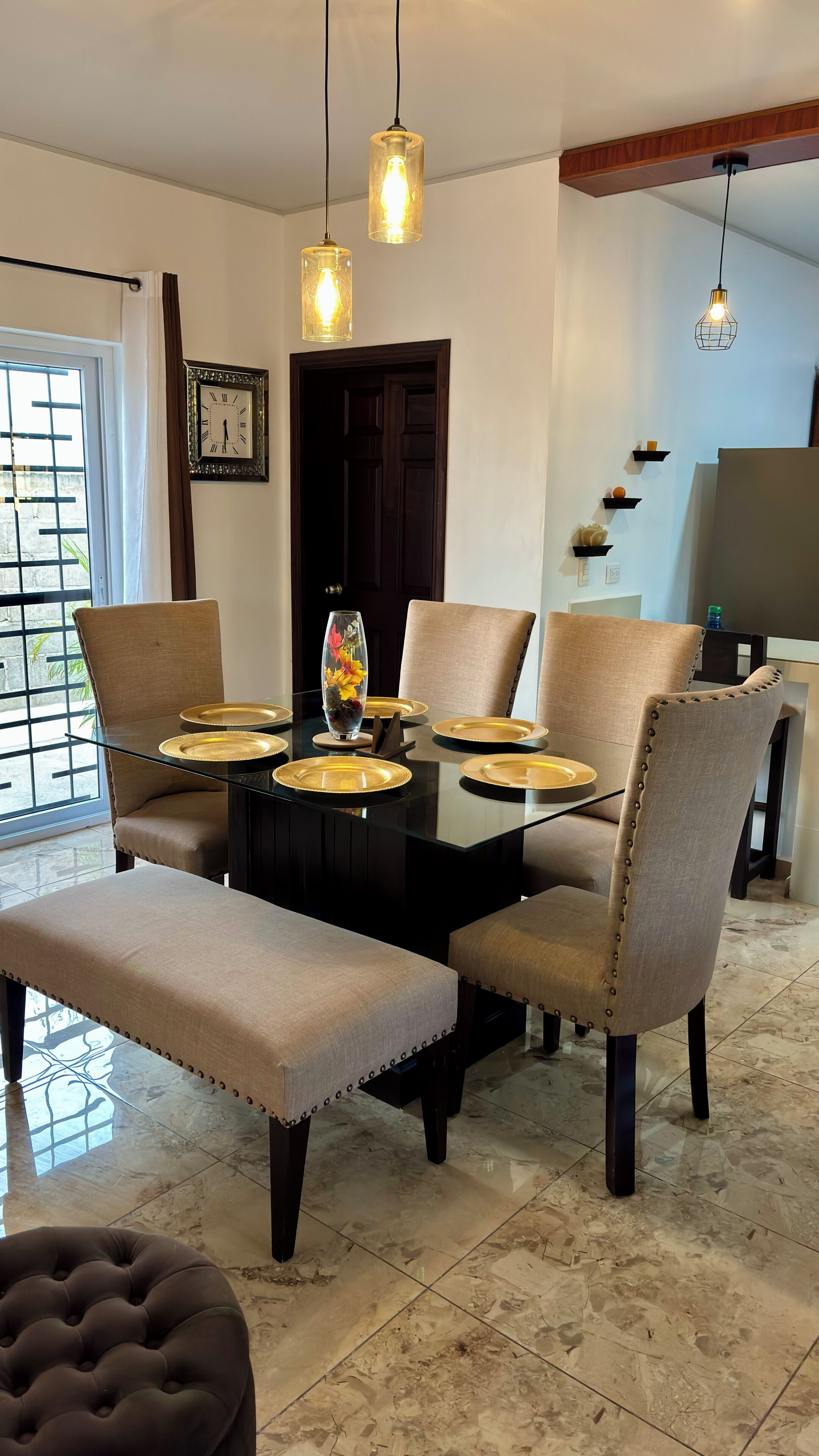 Un comedor con una mesa de vidrio, sillas acolchadas e iluminación colgante.