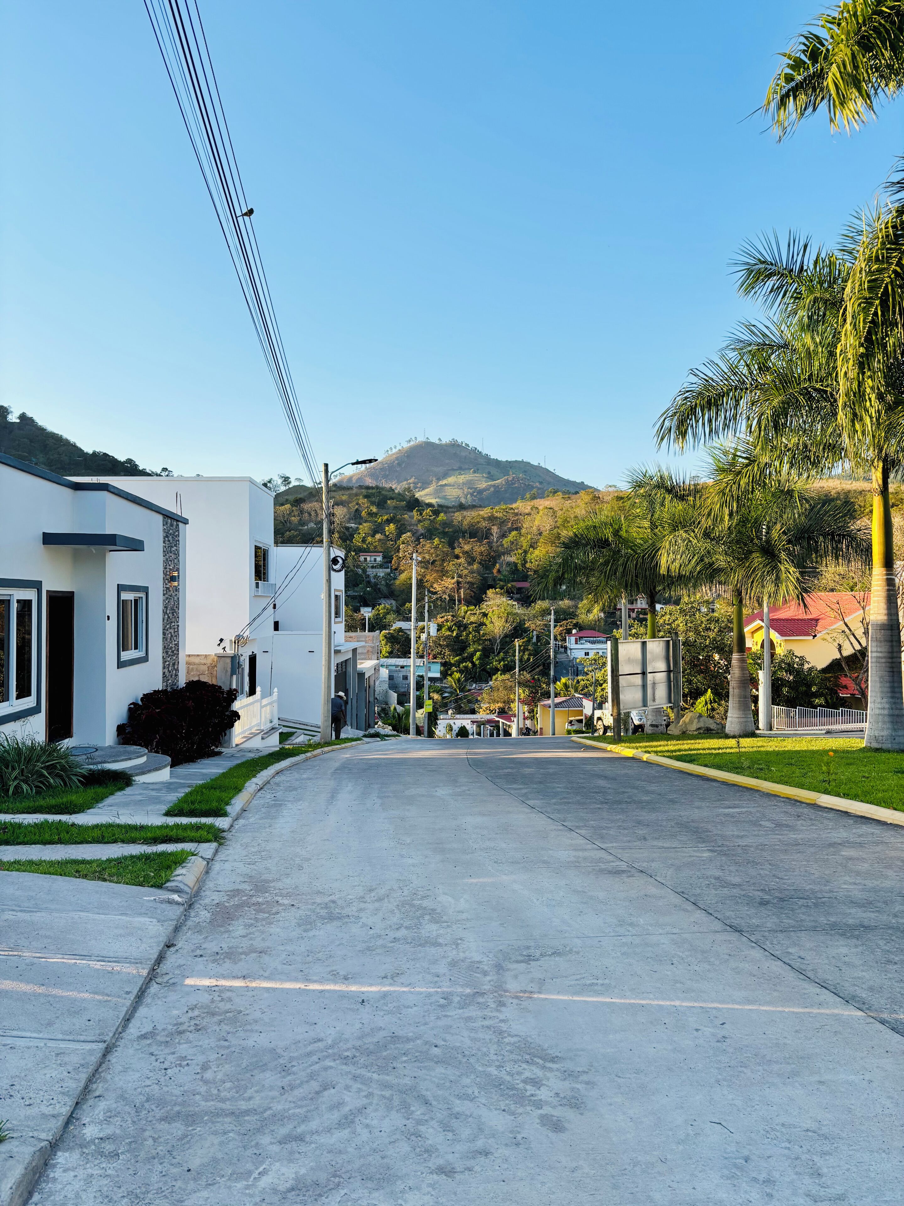 Une rue résidentielle bordée de maisons, un ciel dégagé et un paysage vallonné en arrière-plan.