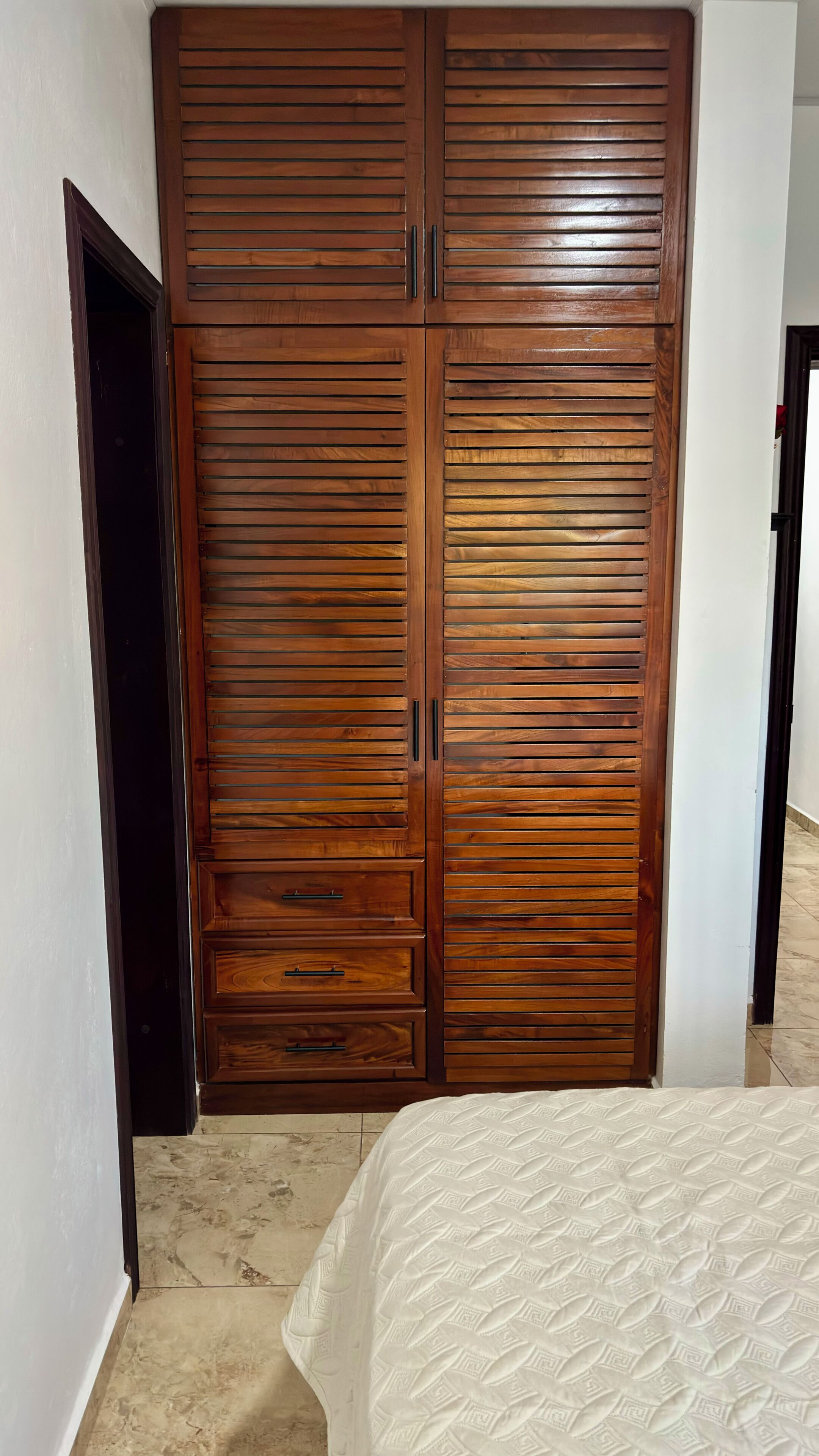 Un dormitorio con un amplio armario de madera, una cama con colcha estampada y suelo de baldosas.