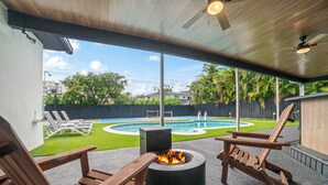 Un patio exterior con sillas de madera, un hogar para fogata y vista a una piscina.