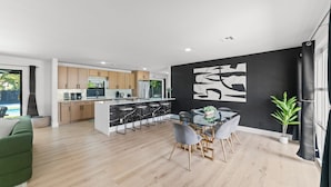 Una cocina moderna con una isla central, un comedor con mesa de vidrio y una pared negra con arte abstracto blanco.