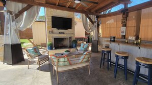 An outdoor patio with a stone fireplace, wicker furniture, a bar area, and a flat-screen TV mounted above the fireplace. - Casa Legacy house — 8 Min Walk to AT&T Stadium, Sleeps 10 (Arlington)