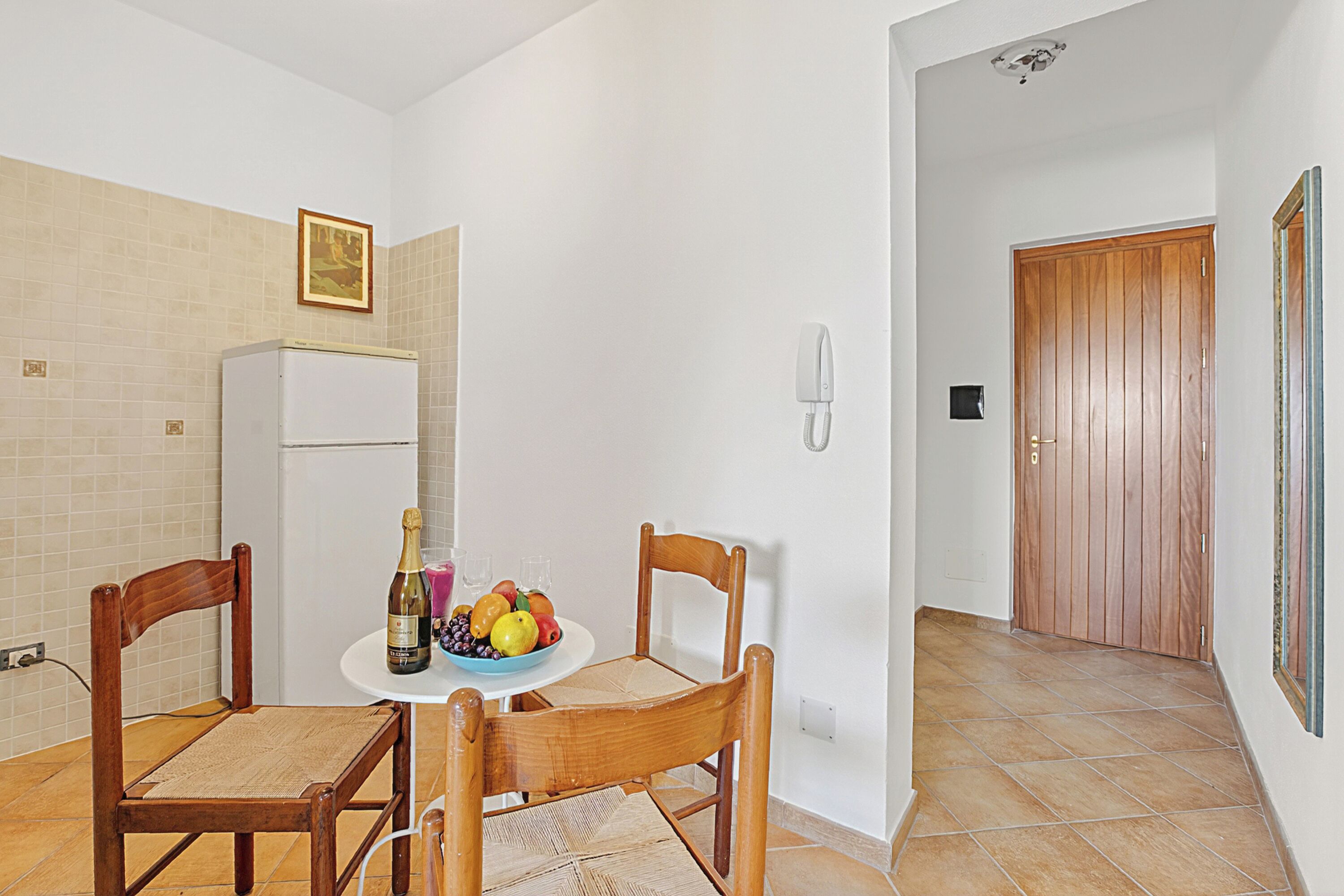Una piccola cucina con un frigorifero, un tavolo di legno con sedie, una ciotola di frutta e una bottiglia di champagne.