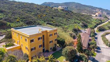 Un edificio giallo a più piani con tetto piatto, circondato da vegetazione e colline sullo sfondo.