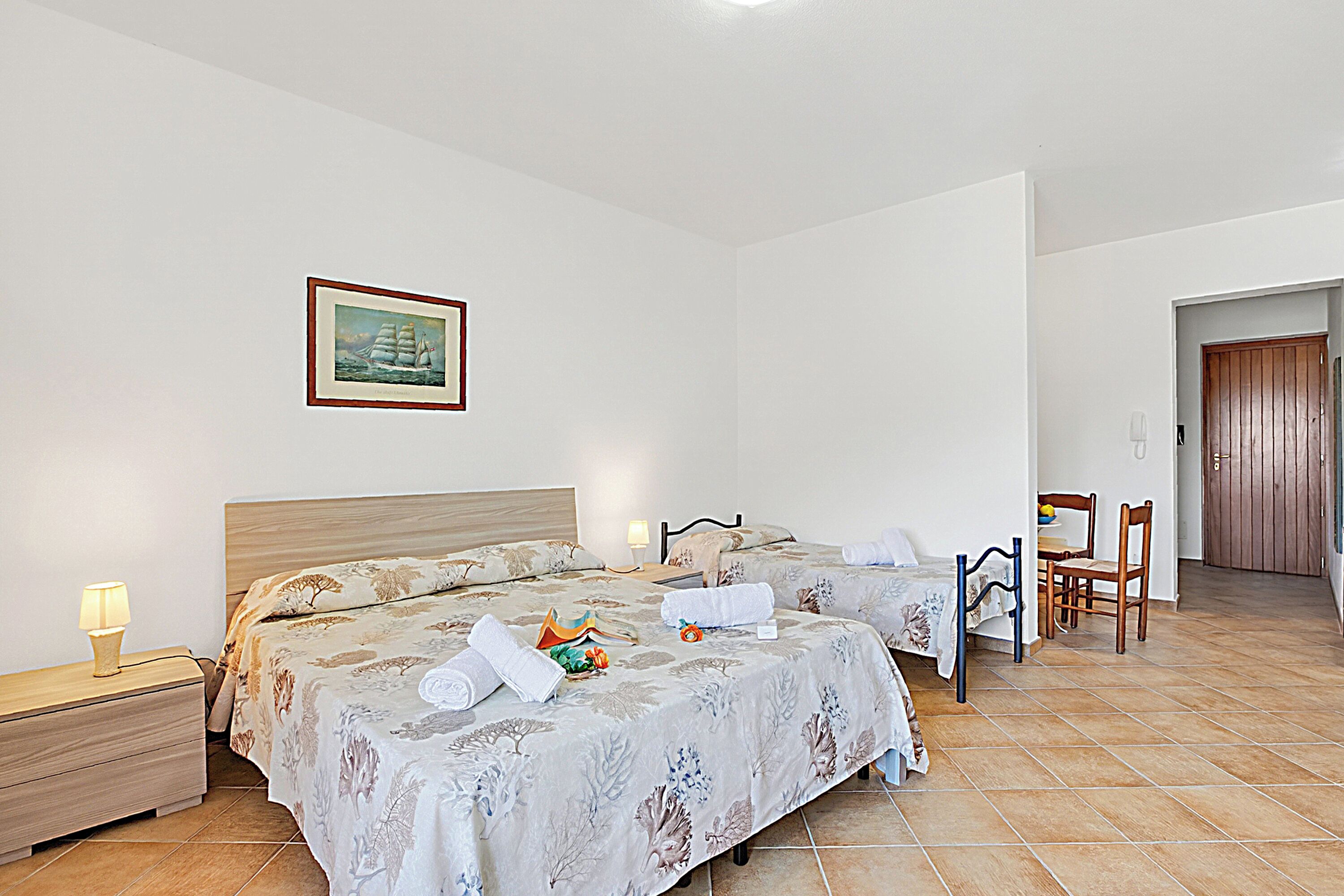 Une chambre d’hôtel avec deux lits, une table de chevet, une chaise, une porte en bois et un tableau encadré au mur.