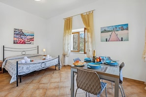 Una habitación con una cama, una mesa de comedor con sillas, un ventanal y dos cuadros en las paredes.