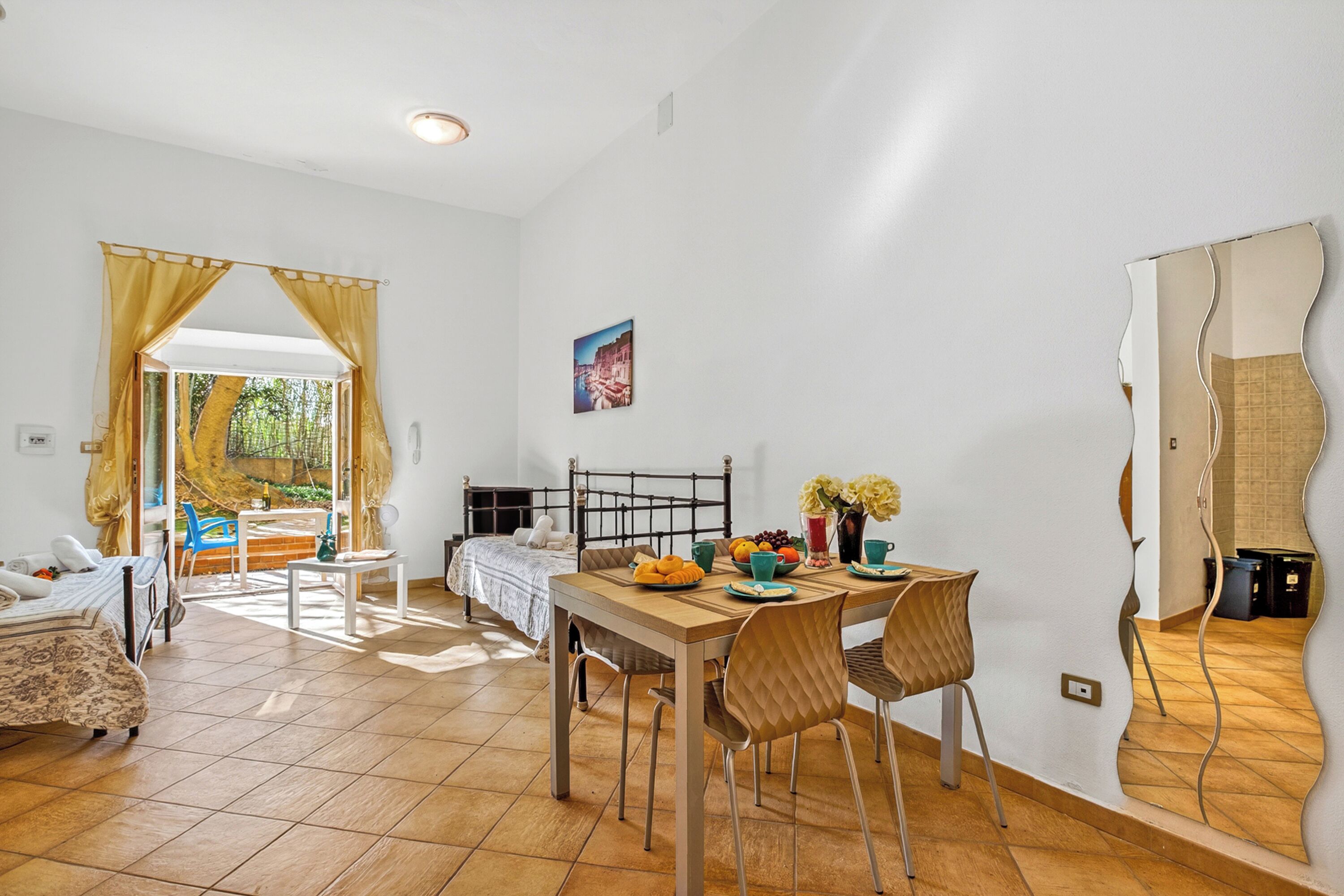 Una habitación con una cama, una mesa de comedor con sillas, una mesita con una silla, un ventanal con cortinas y un cuadro colgado en la pared.