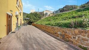 A paved pathway leading to a yellow building with a stone wall and greenery on a hillside. - Residence Argentiera Studio 1 for four, Miniera dell’Argentiera, Italy (Miniera dell’Argentiera)