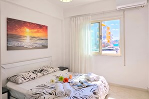A hotel room with a bed, a painting of a sunset, a window with a city view, and a small table with towels and flowers. - Mandalay Suites By The Beach 079, Durres, Albania (Durres)