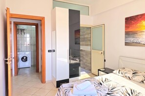 Ein Schlafzimmer mit Bett, Nachttisch, Kleiderschrank und einem Badezimmer inklusive Waschmaschine sowie einem Sonnenuntergangsbild.