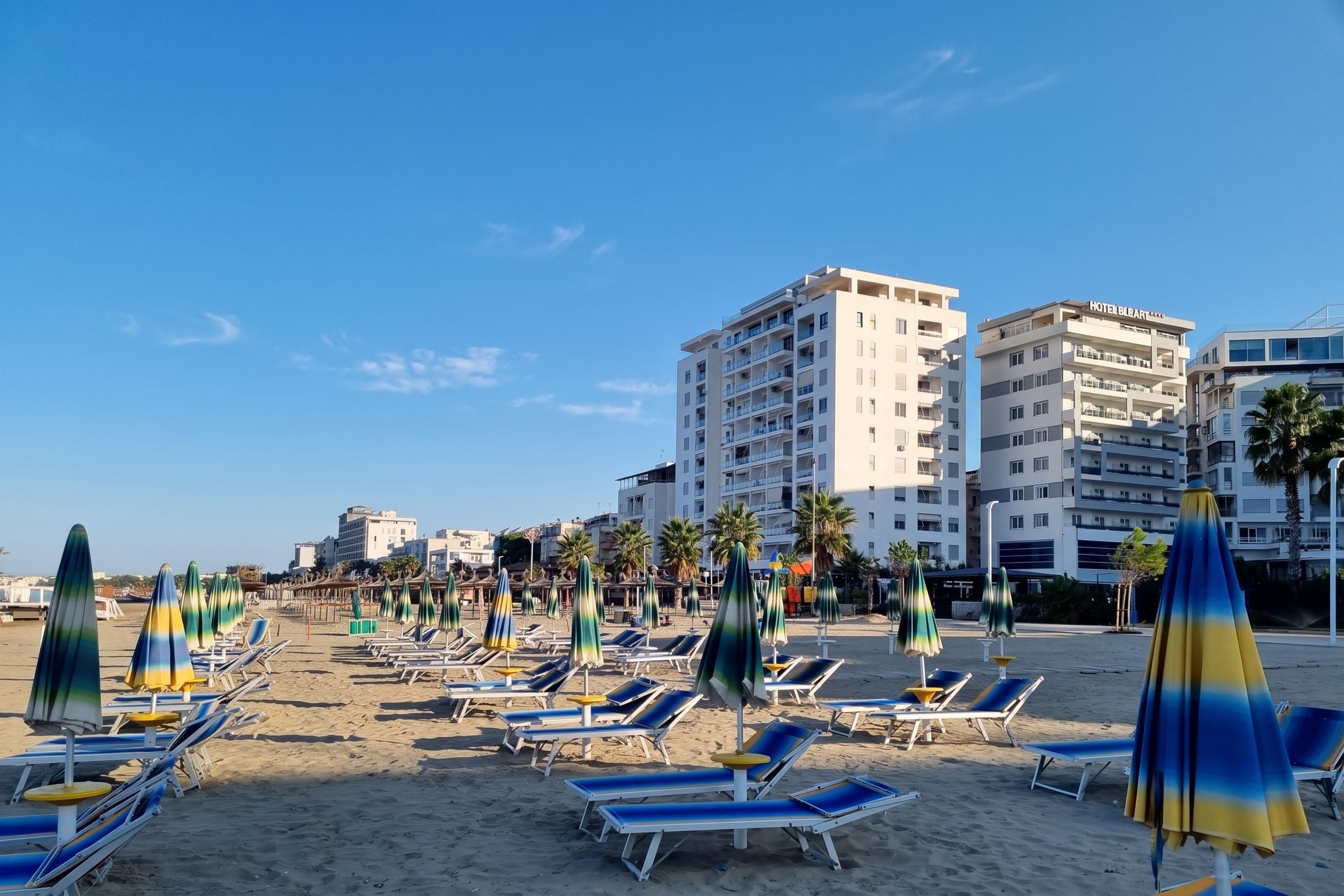 A beach with sun loungers and umbrellas, a hotel in the background, and palm trees.