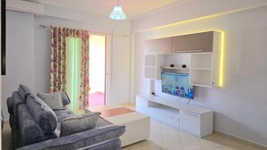 A modern living room with a grey sofa, a flat-screen TV mounted on a white cabinet, and a ceiling light fixture. - Mandalay Suites By The Beach 079, Durres, Albania (Durres)