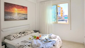 A hotel room with a bed, a painting of a sunset, a window with a view of buildings, and a nightstand with a lamp. - Mandalay Suites By The Beach 079, Durres, Albania (Durres)