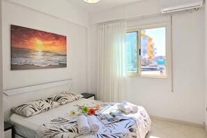 Ein Hotelzimmer mit einem Bett, einem Sonnenuntergangsgemälde, einem Fenster mit Blick auf Gebäude und einem Nachttisch mit einer Lampe.