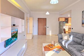 A modern living room with a flat-screen TV, a grey sofa, a coffee table with a tray of muffins, and a wooden door leading to another room. - Mandalay Suites By The Beach 079, Durres, Albania (Durres)