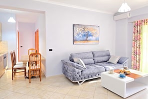 A modern living room with a grey sofa, a dining table with chairs, a painting on the wall, and a window with curtains. - Mandalay Suites By The Beach 079, Durres, Albania (Durres)
