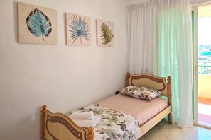 A single bed with a wooden headboard, a floral bedspread, and a pillow. There are three wall-mounted artworks depicting leaves. A white curtain partially covers a window. - Mandalay Suites By The Beach 079, Durres, Albania (Durres)