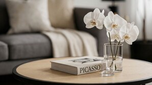 Ein runder Holztisch mit einem Buch mit dem Titel 'PICASSO' und zwei klaren Vasen mit weißen Blumen.