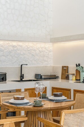 A dining area with a round wooden table, chairs, and a kitchen in the background. - Mareo Villa (Pecatu)