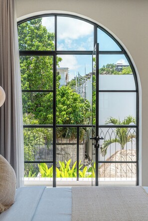 A room with a large arched window, a bed with white bedding, and a view of greenery and buildings outside. - Mareo Villa (Pecatu)