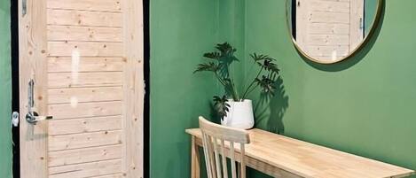 Una habitación con puerta de madera, un escritorio con silla, una planta en maceta y un espejo redondo en la pared.