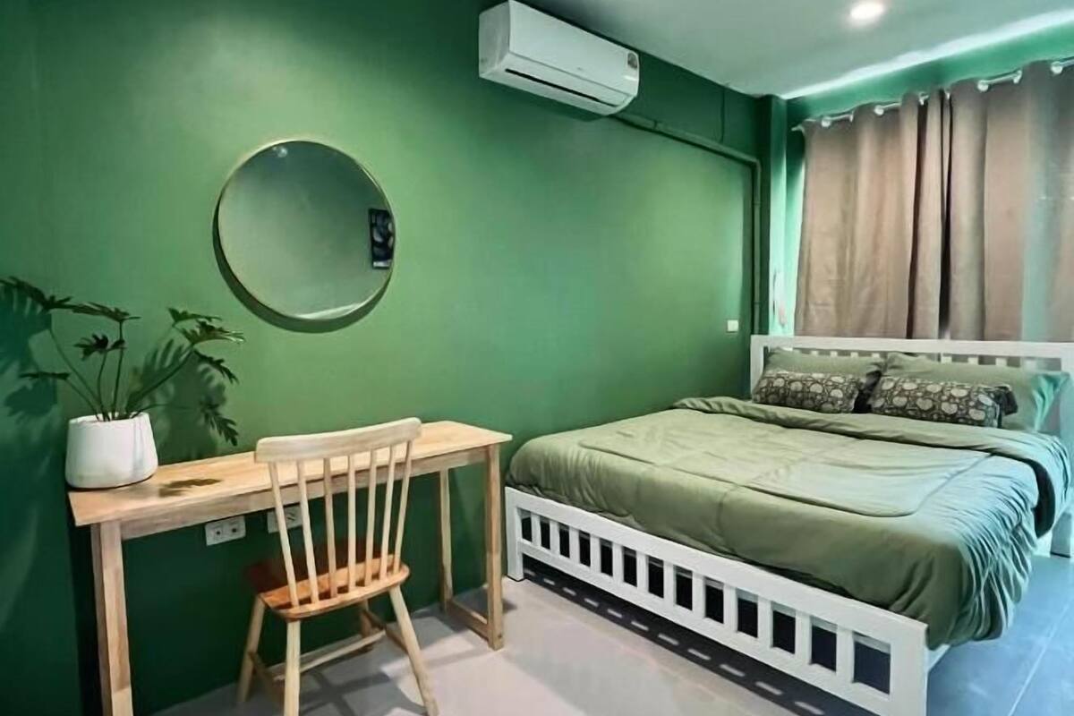 Ένα υπνοδωμάτιο με ένα κρεβάτι, ένα γραφείο, μια καρέκλα, ένα φυτό σε γλάστρα, ένα κλιματιστικό τοποθετημένο στον τοίχο και ένα παράθυρο με κουρτίνες.