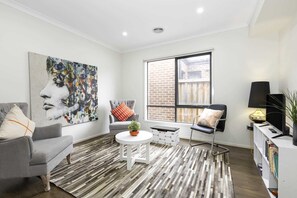 A modern living room with a grey sofa, a round coffee table, a black leather chair, a large abstract painting, a window with a brick wall view, and a bookshelf with books.
