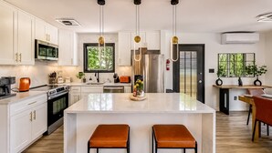 A modern kitchen with white cabinetry, stainless steel appliances, a central island with stools, pendant lighting, and a view of greenery outside. - Lakefront Modern Escape with Hot Tub (East Stroudsburg)