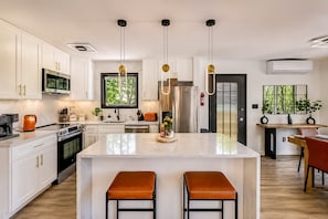 Cocina moderna con muebles blancos, electrodomésticos de acero inoxidable, una isla central con taburetes, iluminación colgante y vistas a zonas verdes.