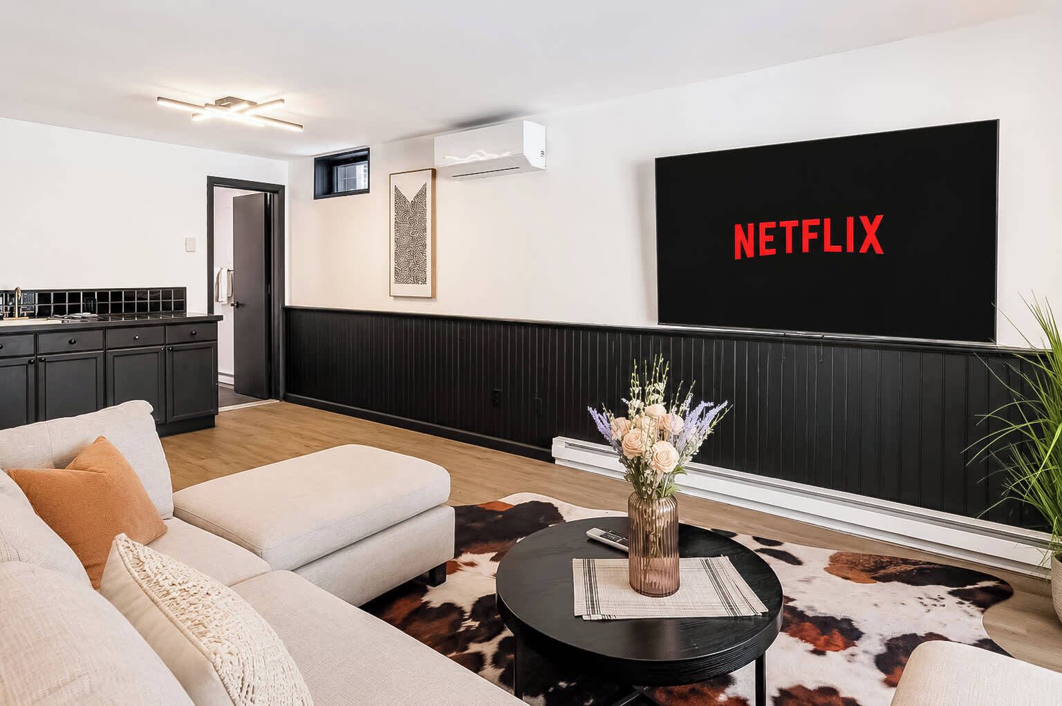 A modern living room with a sofa, a coffee table, a TV wall-mounted above a black and white wall, and a kitchen in the background.