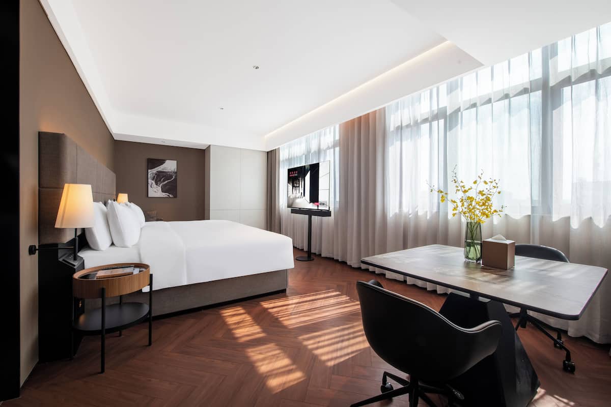 Una habitación de hotel con cama, mesita de noche, lámpara, sillón, escritorio y ventana con cortinas.