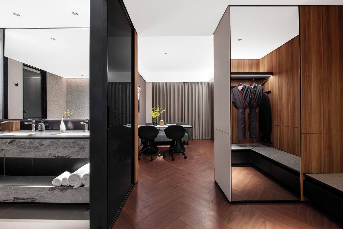 Una habitación de hotel moderna con suelo de madera, tocador con azulejos de mármol, zona de comedor con mesa y sillas, y vestidor con ropa colgada.
